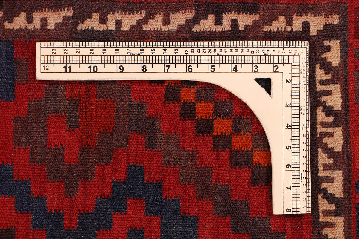 Red kilim rug with dark geometric motifs and measuring tape along edge.