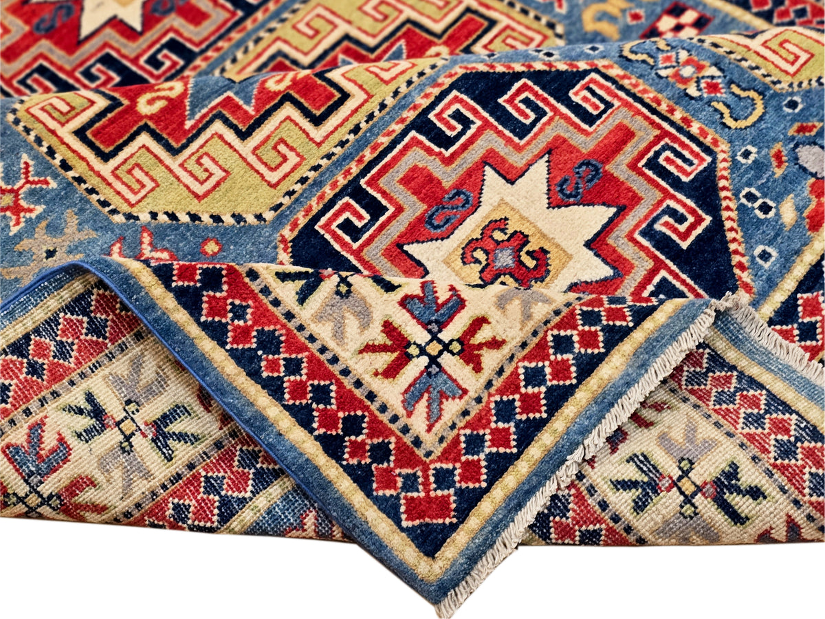 Patterned Kazak rug corner with folded edge, blue ground, and red diamond motifs.